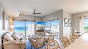 A spacious living room with a large window offering a view of the ocean and cityscape, a ceiling fan, and a dining area with wicker chairs. - Gulf Views ~ Lockout Unit ~ Private Foyer ~ Balcony (Miramar Beach)