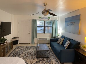 A living room with a ceiling fan, a TV, a couch, a coffee table, a rug, and a window with curtains. - A Nice Place To Stay! (Tybee Island)