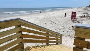 Eine Holztreppe führt zu einem Strand, wo ein Rettungsschwimmerstuhl mit der Aufschrift '419' steht.