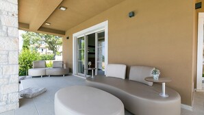 Ein moderner Außenbereich zum Sitzen mit einer gebogenen Couch und Hocker, einem kleinen runden Tisch und einer Glastür, die zu einer überdachten Terrasse führt.