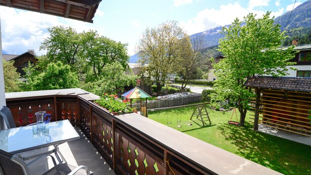 A balcony with a glass table and chairs, a wooden fence, a swing set, and a view of a village with houses and mountains in the background. - Ferienwohnung 50 m² (Kötschach-Mauthen)
