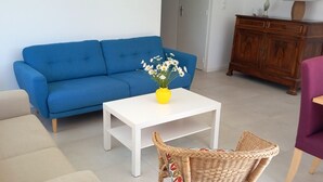 Un salon avec un canapé bleu, une table basse blanche, un fauteuil en osier et un fauteuil violet.