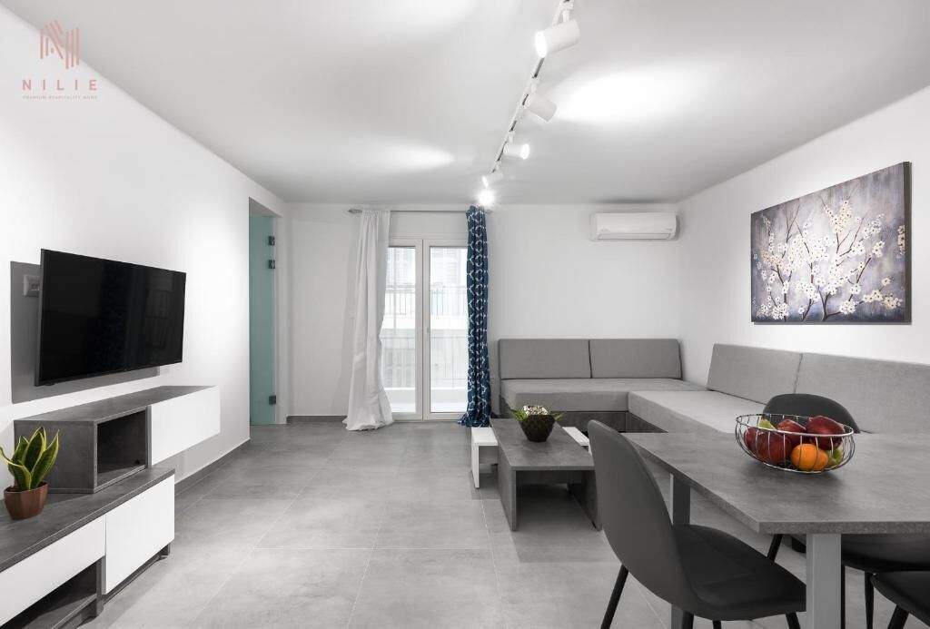 Un salon moderne avec un téléviseur à écran plat, un canapé gris, une table à manger avec des chaises et un panier de fruits.