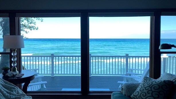 Habitación con una ventana grande que da a una playa y al océano. Hay una cama con un cojín y una lámpara sobre una mesita de noche.