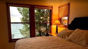 Un dormitorio con un ventanal grande que ofrece vistas al mar y a los árboles, una cama con colcha estampada y una lámpara de noche.