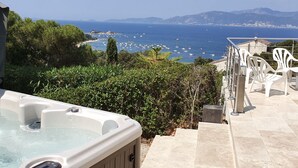 Un jacuzzi con vista despejada al mar y barcos, rodeado de vegetación y muebles de exterior blancos.