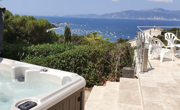 Un jacuzzi con vista despejada al mar y barcos, rodeado de vegetación y muebles de exterior blancos.