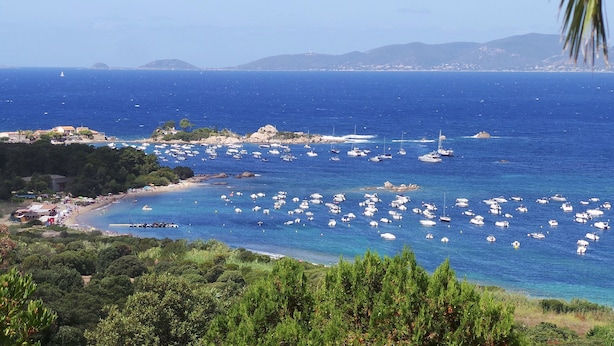 Una zona costera con numerosas embarcaciones fonqueadas en el agua, una playa con sombrillas y montañas al fondo.