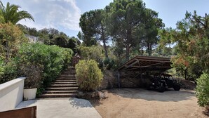 Una escalera de concreto que sube hacia un muro de piedra, rodeada de abundante vegetación y un vehículo todoterreno estacionado bajo una estructura metálica.