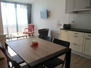 Un comedor con una mesa de madera, sillas negras, una silla roja, una pequeña mesa con una vela, un televisor montado en la pared y una kitchenette con armarios y un microondas.