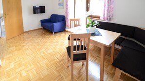 A room with a wooden parquet floor, a blue sofa, a black sofa, a wooden dining table with a chair, a potted plant, and a flat-screen TV mounted on the wall. - Ferienwohnung 35m² (Kötschach-Mauthen)