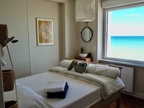 A neatly made bed with pillows and a folded towel, a bedside table with a lamp, a framed artwork on the wall, a round mirror, and a window with a view of the sea. - Maison Spacieuse Front de mer (Quinéville)