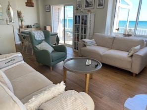 A living room with a sofa, armchair, and a coffee table. There is a view of the sea through large windows. - Maison Spacieuse Front de mer (Quinéville)