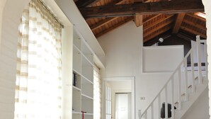 A spacious living area with a high ceiling, wooden beams, a staircase, and a bookshelf.