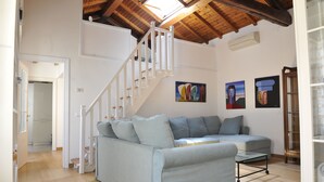 A living room with a light blue sofa, a glass coffee table, and a wooden staircase. There are abstract paintings on the wall and a door to the right.