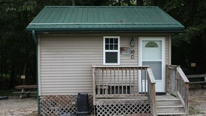 Une petite cabane à un seul étage, avec un toit vert, une porte d’entrée et un petit porche doté d’une balustrade en bois.