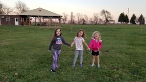 Trois enfants se tenant la main dans un champ herbeux, avec une grange et des arbres en arrière-plan.