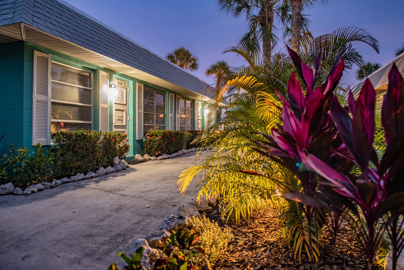 A two-story building with a teal exterior and white shutters, surrounded by tropical plants and a paved driveway.