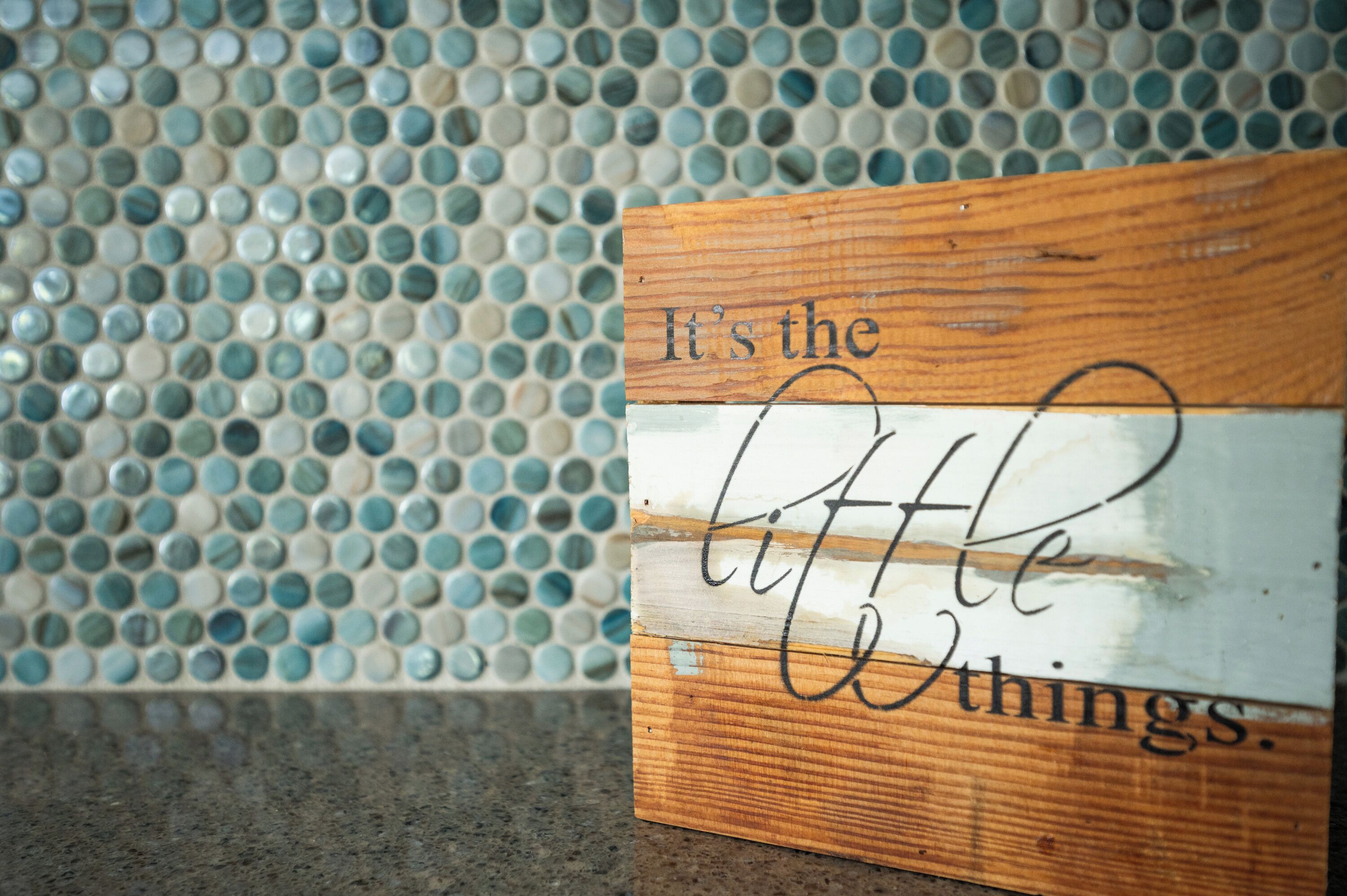 Un cartello di legno con la scritta "It's the little things" davanti a un muro rivestito di mosaici.