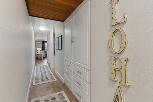 A hallway with a wooden ceiling, white walls, and a patterned rug. There are framed pictures on the wall and a white cabinet with drawers. - Updated Kamaole Sands Condo (Kihei)