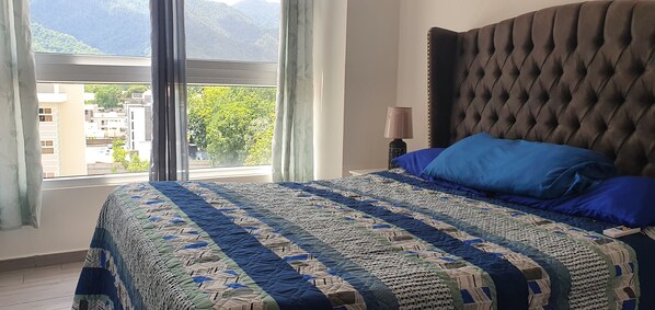 A bedroom with a large bed, a tufted headboard, and a view of buildings and trees through a window. - Exclusive Apartment Arboleda 186 (San Pedro Sula)
