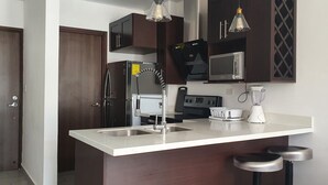 A modern kitchen with dark wooden cabinets, stainless steel appliances, a white countertop, and two bar stools. - Exclusive Apartment Arboleda 186 (San Pedro Sula)