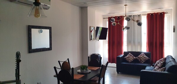 Ein Wohnzimmer mit einer Couch, einem Esstisch mit Stühlen, einem Fernseher und einem roten Vorhang.