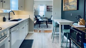 A modern kitchen with white cabinets, a black faucet, and a microwave. There is a dining area with a table and chairs, and a painting on the wall.
