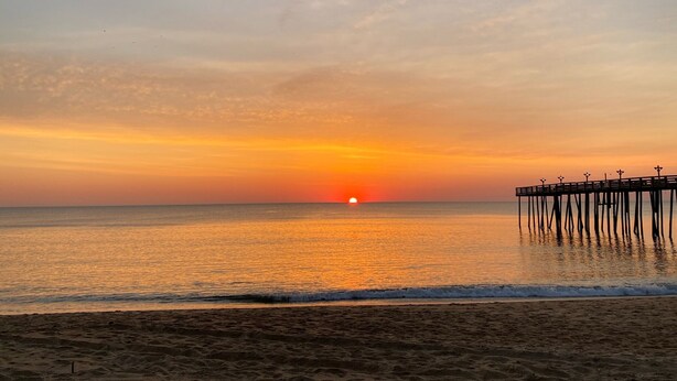 A beach with a pier extending into the water, a sunset in the background, and footprints in the sand.