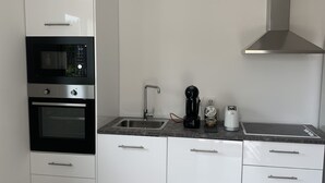 Une cuisine moderne avec des meubles blancs, un four encastré, une hotte de cuisinière en acier inoxydable, un évier et un plan de travail sur lequel se trouvent une machine à café et un grille-pain.
