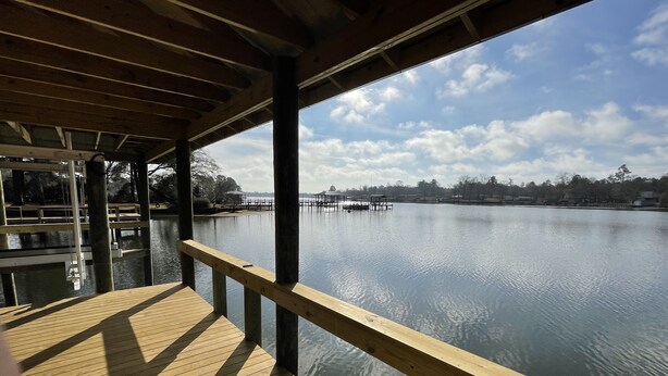 Una terraza de madera con barandilla que da a un lago tranquilo, con muelles y edificios al fondo.