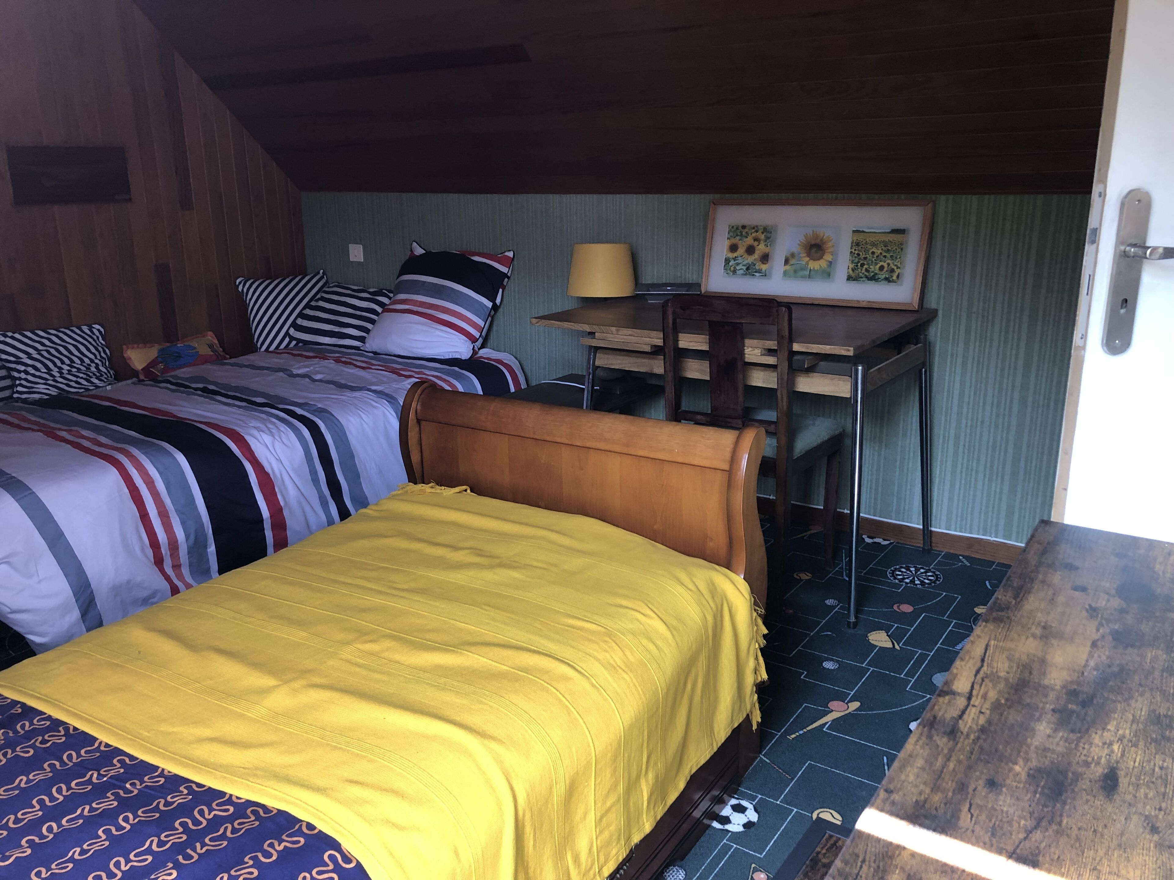 A room with two beds, one with a yellow bedspread and the other with striped bedding. There is a desk with a lamp and a framed picture of sunflowers on the wall.