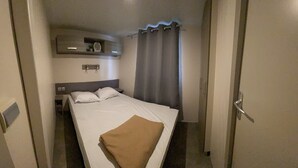 Hotellihuone, jossa on sänky, yöpöytä, seinälle kiinnitetty hylly, jossa on kello, ikkuna verhoilla ja ovi.