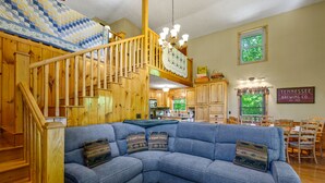 A cozy living room with a blue sectional sofa, wooden staircase, and a kitchen area in the background. - Beautiful Amazing Setting, Fire Pit & Pool Table, Large Wrap Around Deck w/ Spa! (Sevierville)