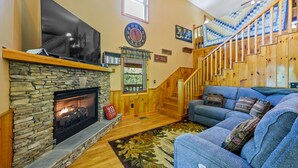 A cozy living room with a stone fireplace, a large flat-screen TV, a blue sofa, and a wooden staircase. - Beautiful Amazing Setting, Fire Pit & Pool Table, Large Wrap Around Deck w/ Spa! (Sevierville)