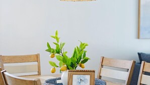 A dining area with a wooden table, four chairs, a woven pendant light, a vase with green leaves and yellow fruits, and a framed picture. - Beachfront 3BR Condo with Oversized Gulf View Balcony, Pool and Spa (Indian Shores)