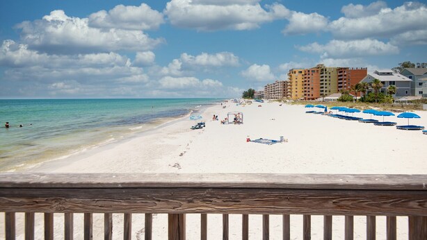 A beach with white sand, a clear blue sky, and a few clouds. There are blue umbrellas and a wooden fence in the foreground. - Beachfront 3BR Condo with Oversized Gulf View Balcony, Pool and Spa (Indian Shores)