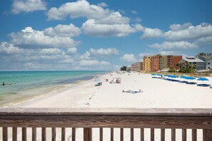 Une plage de sable blanc, un ciel bleu clair et quelques nuages. On aperçoit des parasols bleus et une clôture en bois au premier plan.