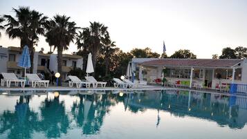 A swimming pool with lounge chairs and umbrellas, surrounded by palm trees and a building with a tiled roof.