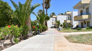 A pathway lined with palm trees and red flowers, leading to a building with balconies and a blue railing. - 1-bedroom Kos Olympia Apartment at Lambi Beach (Kos)