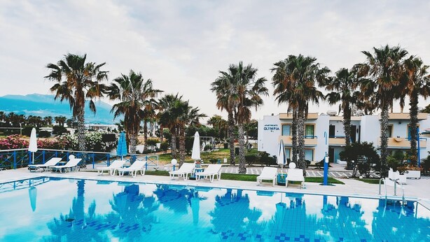 A swimming pool with lounge chairs and palm trees, surrounded by a building labeled "Apartments Olympia." - 1-bedroom Kos Olympia Apartment at Lambi Beach (Kos)