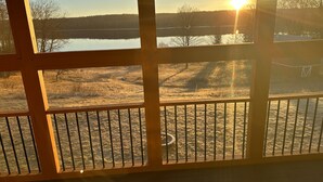 Une vue à travers une fenêtre à cadre en bois, donnant sur un lac et une zone herbeuse, avec un coucher de soleil en arrière-plan.