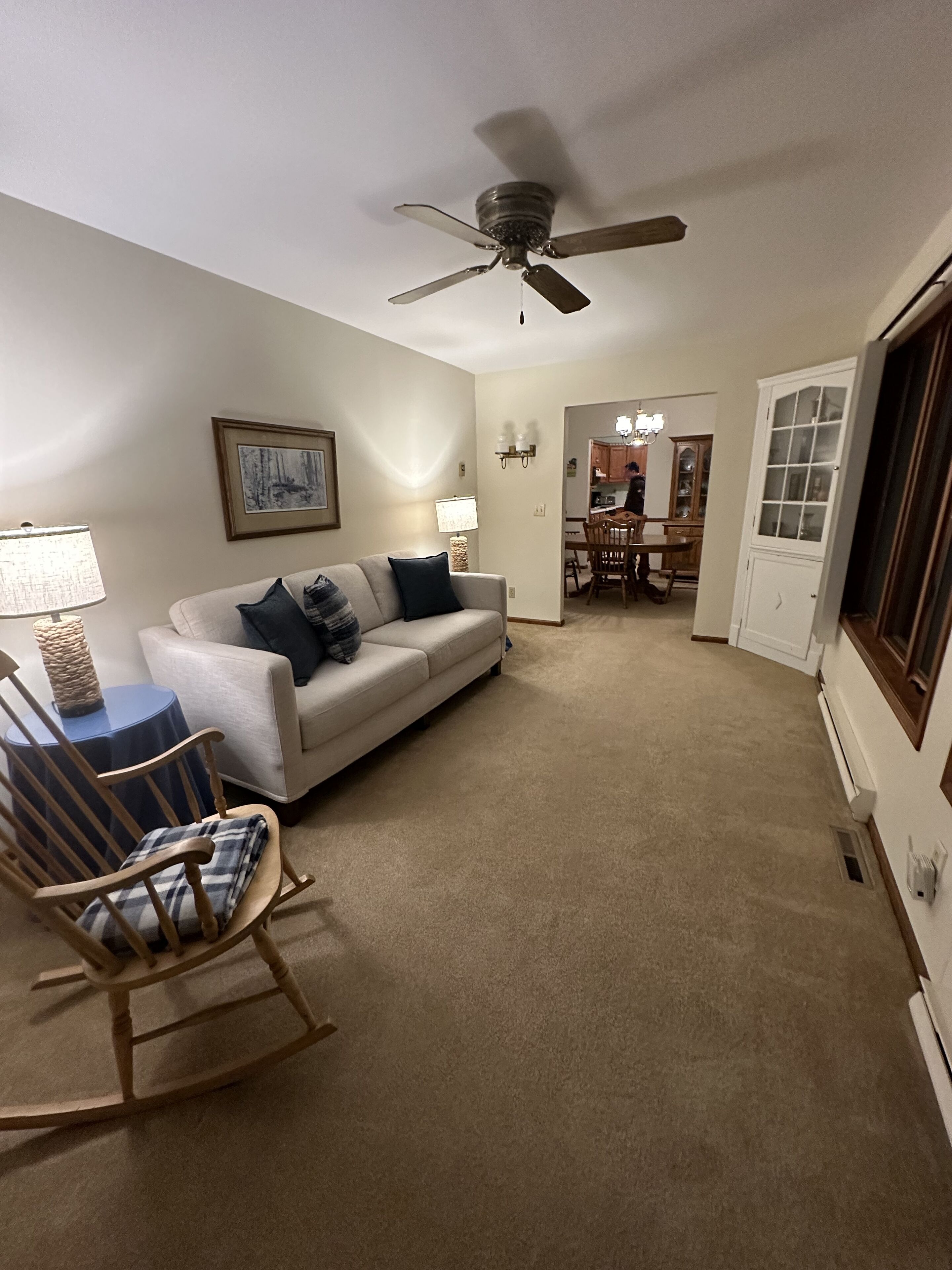 A living room with a beige carpet, a white sofa with dark cushions, a wooden rocking chair, a small blue table, and a ceiling fan.