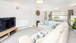 A modern living room with a flat-screen TV, a sofa with patterned cushions, a fireplace, and a large window with curtains. - Property 5 Minutes Walk From Trearddur Bay Beach (Holyhead)