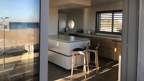 A modern kitchen with a central island, two bar stools, a window with blinds, and a view of the beach. - Seafront -  feet in the sand - the most beautiful view of Narbonne Plage (Narbonne Plage)