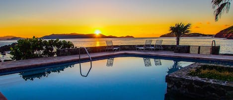 A pool with lounge chairs and a sunset view.