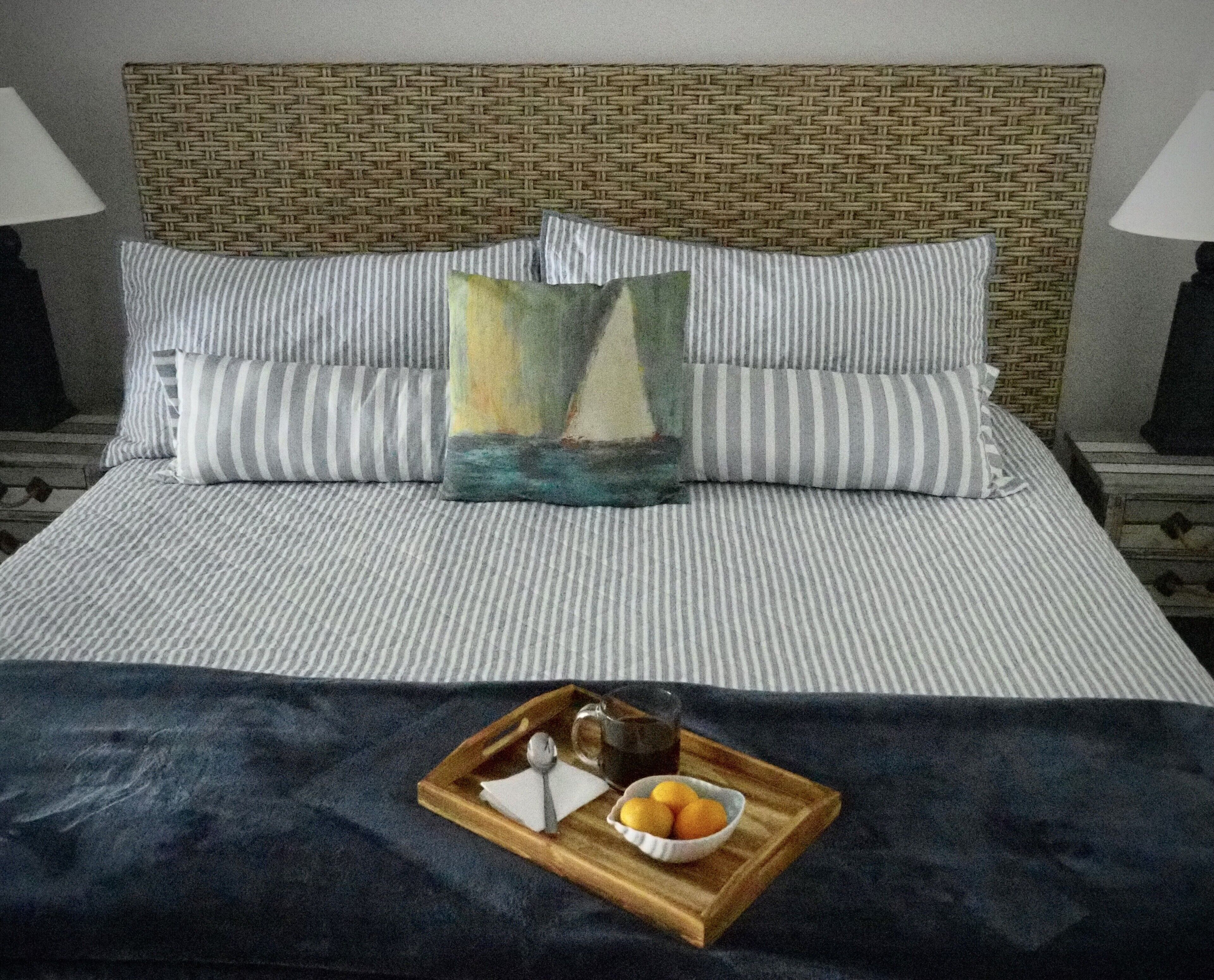 Un lit bien fait, avec une literie à rayures, un coussin décoratif et un plateau sur lequel se trouvent un verre et quelques oranges.
