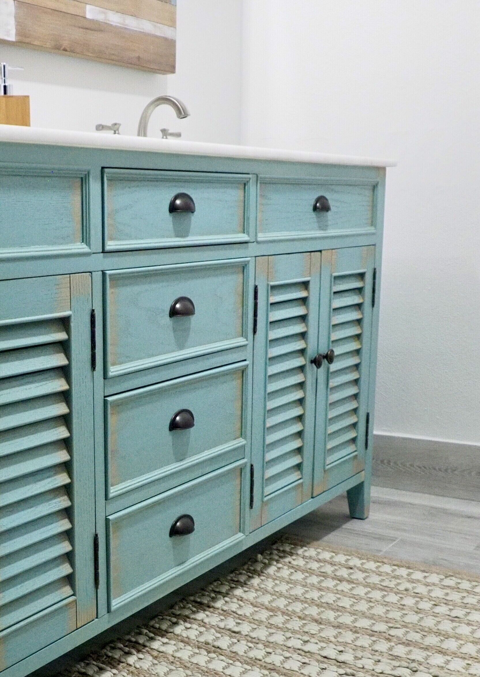 A teal-colored wooden cabinet with drawers and louvered doors, a white countertop above, and a textured rug on the floor.