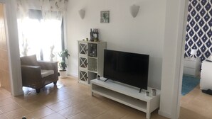 A living room with a flat-screen TV on a white stand, a glass cabinet with bottles, a wicker sofa, and a potted plant. - Refúgio Ls (Guia)