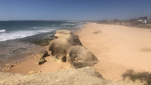 A beach with clear skies, rocky formations, and a distant shoreline with buildings. - Refúgio Ls (Guia)